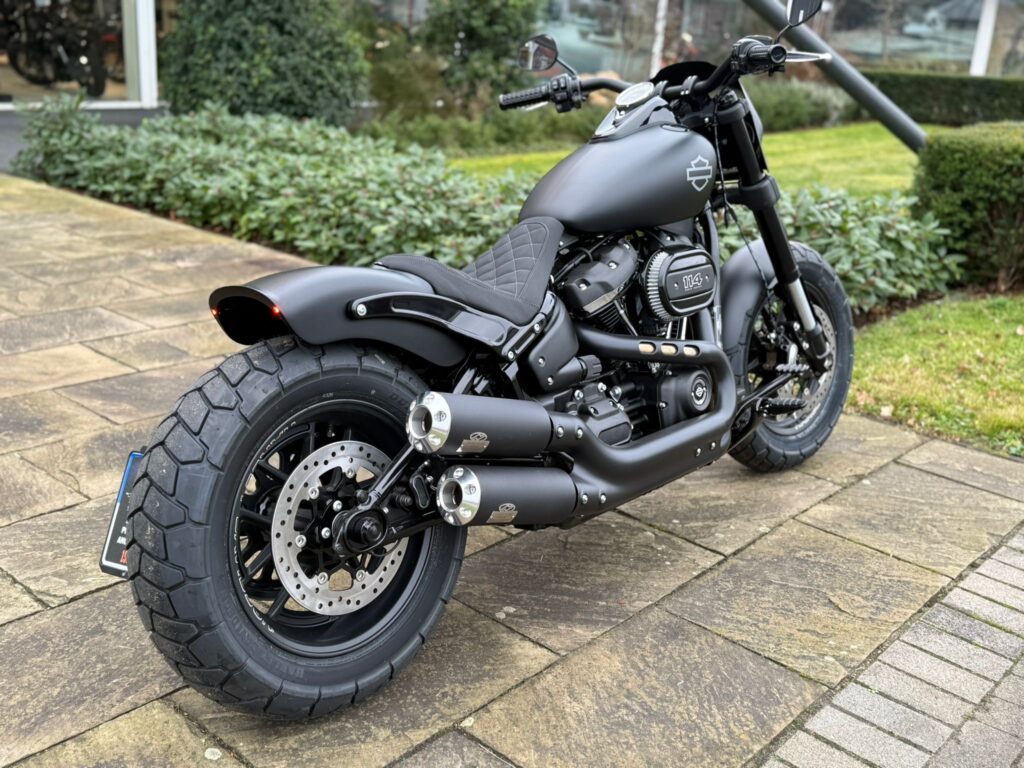Eine matte Harley-Davidson-Motorrad mit einem auffälligen Reifenprofil und sportlichen Auspuffanlagen steht auf einem gepflasterten Weg vor einer grünen Hecke.
