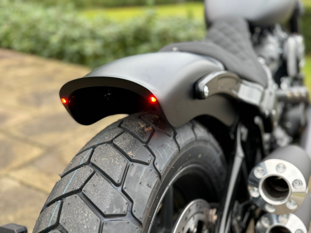 Detailaufnahme des Hinterrads einer Harley-Davidson mit schwarzem Schutzblech und roten Rücklichtern auf einem gepflasterten Boden.