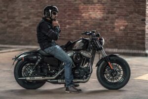 Ein Motorradfahrer in Schutzkleidung und Helm sitzt auf einer schwarzen Harley-Davidson in einer urbanen Umgebung.