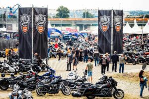 Blick auf eine lebhafte Harley-Davidson-Veranstaltung mit zahlreichen Motorrädern, Banner und Besuchern in einem Outdoor-Bereich.
