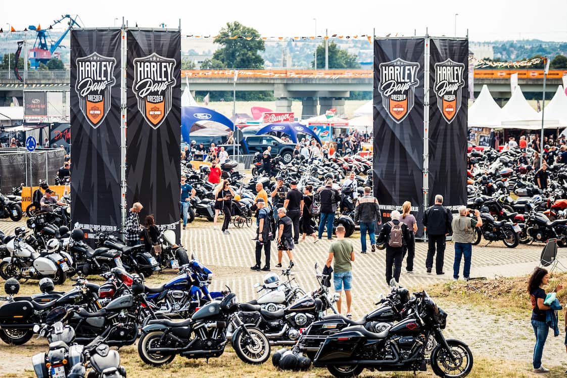 Blick auf eine lebhafte Harley-Davidson-Veranstaltung mit zahlreichen Motorrädern, Banner und Besuchern in einem Outdoor-Bereich.
