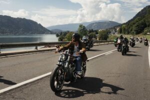 Motorradfahrer auf einer Harley-Davidson fährt entlang einer malerischen Straße am Wasser mit anderen Motorrädern im Hintergrund.