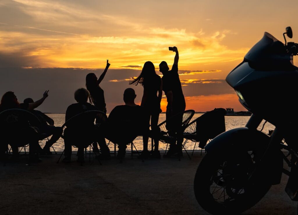 Eine Gruppe von Menschen genießt den Sonnenuntergang am Wasser, während eine Harley-Davidson-Motorrad im Vordergrund steht.