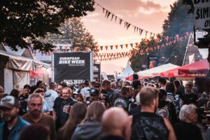 Eine lebhafte Menschenmenge feiert die European Bike Week am Faak am See mit bunten Fahnen und Ständen, die Motorrad-Zubehör und Bekleidung anbieten.