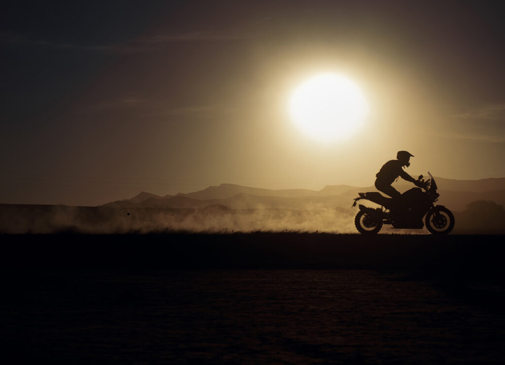 Ein Biker fährt im Sonnenuntergang auf einem Motorrad durch eine staubige Landschaft.