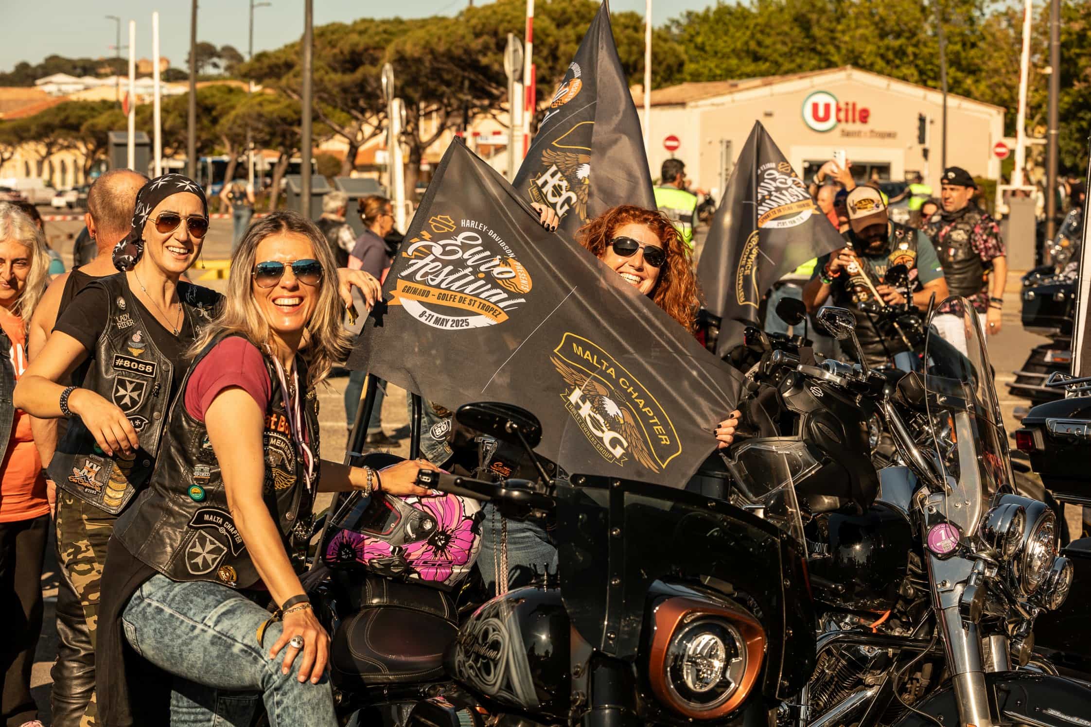 Verschiedene Motorradfahrerinnen und Motorradfahrer feiern zusammen mit Harley-Davidson Flaggen und Motorrädern auf einem Festival.