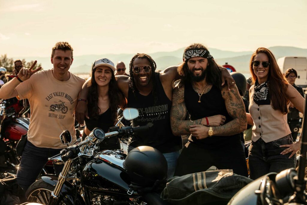 Fünf Motorradliebhaber posieren lächelnd vor ihren Harley-Davidson Bikes bei einem Event im Freien.