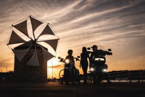 Zwei Motorradfahrer stehen bei Sonnenuntergang vor einem historischen Windmühlengebäude und genießen die Atmosphäre des Abends.