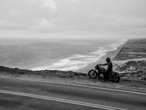 Ein Motorradfahrer genießt die Aussicht auf eine Küstenstraße und den Ozean in einer schwarz-weißen Szenerie.