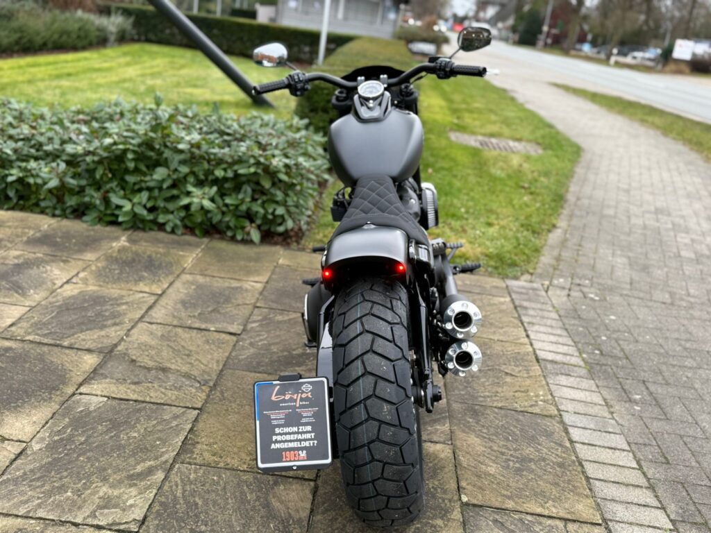 Eine schwarze Harley-Davidson mit markantem Hinterrad und sportlichem Design steht auf einem gepflasterten Weg vor einem grünen Hintergrund.